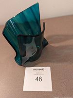 Photo of abstract art glass vessel showing its dark blue-green coloration and asymmetrical sculptural form on a neutral surface.