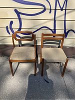 Full view of a pair of teak mid-century modern chairs showing front and seats, set on pavement with graffiti background.