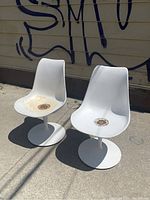Two white tulip chairs outdoors, showing front and top of seats, with rust and discoloration on one seat.