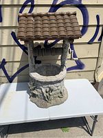 Front view of a rustic garden wishing well on a white table with a brown tiled roof, faux wood supports, and textured stone base