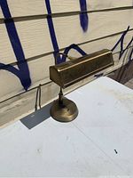 Full side view of one brushed gold tone bankers style desk lamp with adjustable goose neck and vented rectangular metal shade on round base.