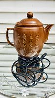 Photo of traditional Chinese-style brown ceramic electric tea kettle with Chinese characters around the body and attached warming base with electric cord in front of a beige wall.