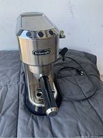 Front angle of the stainless steel DeLonghi espresso coffee maker with attached portafilter and visible electrical cord.