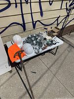 Overall view of lot showing two white hard hats, orange safety vest, rigid pipe conduit bender, and multiple metal junction boxes and electrical components on white folding table.
