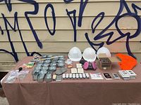 Overview of assorted electrical boxes, outlets, faceplates, three white hard hats, pink respirator, and a box with light fixtures and additional electrical items on a table.