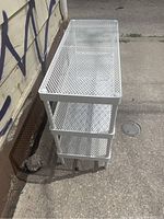 Three-tier white steel mesh shelving unit outdoors against a wall with graffiti, showing no visible blemishes or damage.