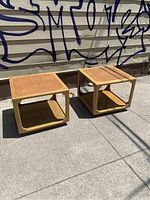 Photo showing the pair of square mid-century modern end tables with leather inlaid tops, light wooden frame, and lower shelf, placed outdoors on concrete against a wall with graffiti.