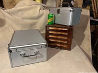 Two silver metal rectangular media storage boxes with ribbed texture and a vintage wooden case with clear orange drawers stacked together with a green soda can for size reference.