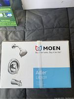 Box front showing Moen Adler L82839 shower set components: showerhead, valve handle, bathtub spout, chrome finish.