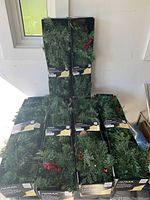 Six boxed Noma brand Preston pre-lit garlands with artificial pine branches and red berry details stacked by a window.