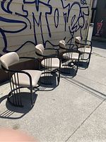 Four post modern dining chairs with black steel frames and beige upholstery, shown outdoors against a wall with graffiti.
