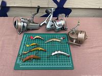 Photo of three vintage fishing reels and seven antique fishing lures laid out on a pink surface with a green measuring mat beneath lures.