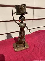 Front view of bronze figure of a woman standing on a wooden base, carrying a basket on her head, with detailed skirt pattern and red bead earrings.