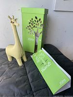 Photo showing synthetic leather beige giraffe item, original green packaging box with tree cutout design, and matching product tag.
