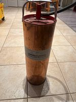 Front angle of tall cylindrical brass fire extinguisher with red pump handle and silver label showing model and instructions