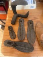 Five iron shoe molds of various sizes and a wooden shoe mold with a handle on a wooden surface.