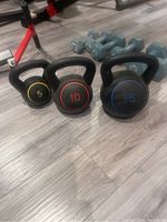Photo shows three kettlebells in 5 lb (yellow), 10 lb (red), and 15 lb (blue) weights with black handles on floor with dumbbells in background.