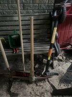 Various wooden-handled garden and yard tools including a pickaxe, rakes, broom, and handheld green seed/fertilizer spreaders on a wooden bench.