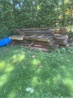 Stacked hardwood slabs of varied widths stored outdoors against hedge, with moss and aged appearance