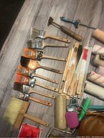 Assorted paint brushes, rollers, wooden handles, and paint scrapers photographed on floor