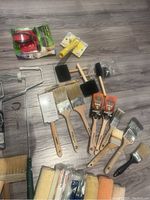 Various paint brushes, foam brushes, some unopened red hand edger, paint can opener tool, and stir sticks arranged on floor.