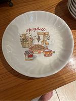 Top view of white ceramic spaghetti bowl with colorful kitchen utensils and food graphic labeled 'Spaghetti'. Scalloped edge design.