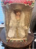Porcelain doll seated in original unopened box, wearing white faux fur coat and gold dress with floral embroidery, brown hair with bangs