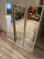 Three identical rectangular mirrors standing upright on a floor with light wood pattern. All have white framing and reflect room contents such as furniture and boxes.
