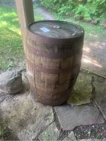 Side view of a large wooden wine barrel under a wooden shelter on natural stone flooring in an outdoor setting with green grass and trees in the background.
