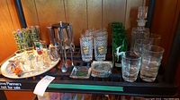 Wide view of barware items on shelf, showing glasses, cocktail tools in stand, decanter, tray, and coasters
