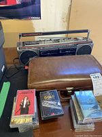 Portable dual cassette stereo unit model 3-5631 A, brown leather carry case, and assortment of cassette tapes on table.