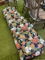 Two thick outdoor cushions stacked, floral pattern, sitting on green carpet with wooden chairs nearby.