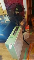 One photo shows the large white box fan in front with the other fans and heater in the background on the floor by a black leather couch and wooden floor.