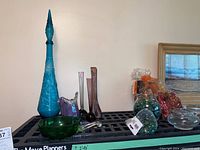 Wide shot showing tall blue vintage glass bottle decanter with stopper, purple and clear bud vases, green blown glass bowl with handle, glass beads in bags, and potpourri.