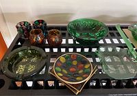 Overview of entire glassware set showing green bowl, amber ashtrays and glasses, green coasters, and tray on black grid shelf.