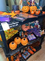 Various ceramic pumpkin decorations, Halloween-themed items including light strings, boxed products, and small decorative pieces arranged on black shelves.