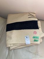 Folded wool blanket on a cream-colored leather surface, showing cream and navy blue stripes and two small green labels.