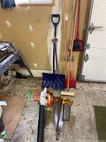 Photo showing the full set of items including the leaf blower, hand saws, brooms, shovel, and chopper arranged near a garage wall.