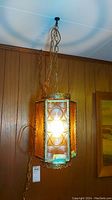 Full view of hanging lamp suspended from ceiling showing glowing amber glass and metal frame with chain and electrical cord.