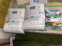 Two packaged Queen size luxury mattress pads from Home By Design, white color, with labels showing product details, placed on a table alongside stacks of folded linen tea towels and dish cloths.