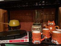 Photo showing yellow enamel fondue pot with wooden handle atop black electric fondue base, Osterizer blender with clear glass pitcher, orange cat-themed ceramic mugs, and defrosting set box stacked underneath.