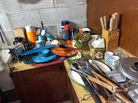 Wide shot of the kitchenware spread on table and counter in basement showing mugs, thermos, serving trays, knives, utensils, and appliance.