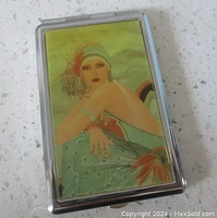 Front view of silver-toned compact mirror showing Art Deco 1920s flapper girl image in green and orange tones.