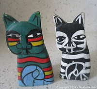 Front view of two Laurel Burch wooden handpainted cat figurines, one dark green with red, yellow, and blue stripes, the other black and white with bold stripes.