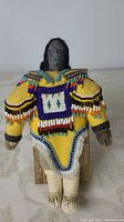 Front view of the doll showing yellow fabric clothing with beadwork and stone head with black hair.