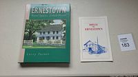 Photo of two items side-by-side: the larger book 'Ernestown Rural Spaces, Urban Places' and smaller pamphlet 'Mills of Ernestown' with a lot number card visible