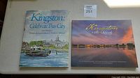 Two books laid flat, showing covers: 'Kingston: Celebrate This City' with watercolor front and 'Kingston & The Islands Then & Now' with sunset photo.