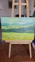 Full view of the oil painting on easel showing the landscape with hills, trees, and fence.