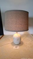 Front full view of the pottery base table lamp fully assembled with fabric shade in neutral tones, lit and plugged in.