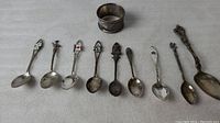 Photo showing full set of 8 souvenir spoons lined up with a silver napkin ring behind them, arranged horizontally on a white textured surface.